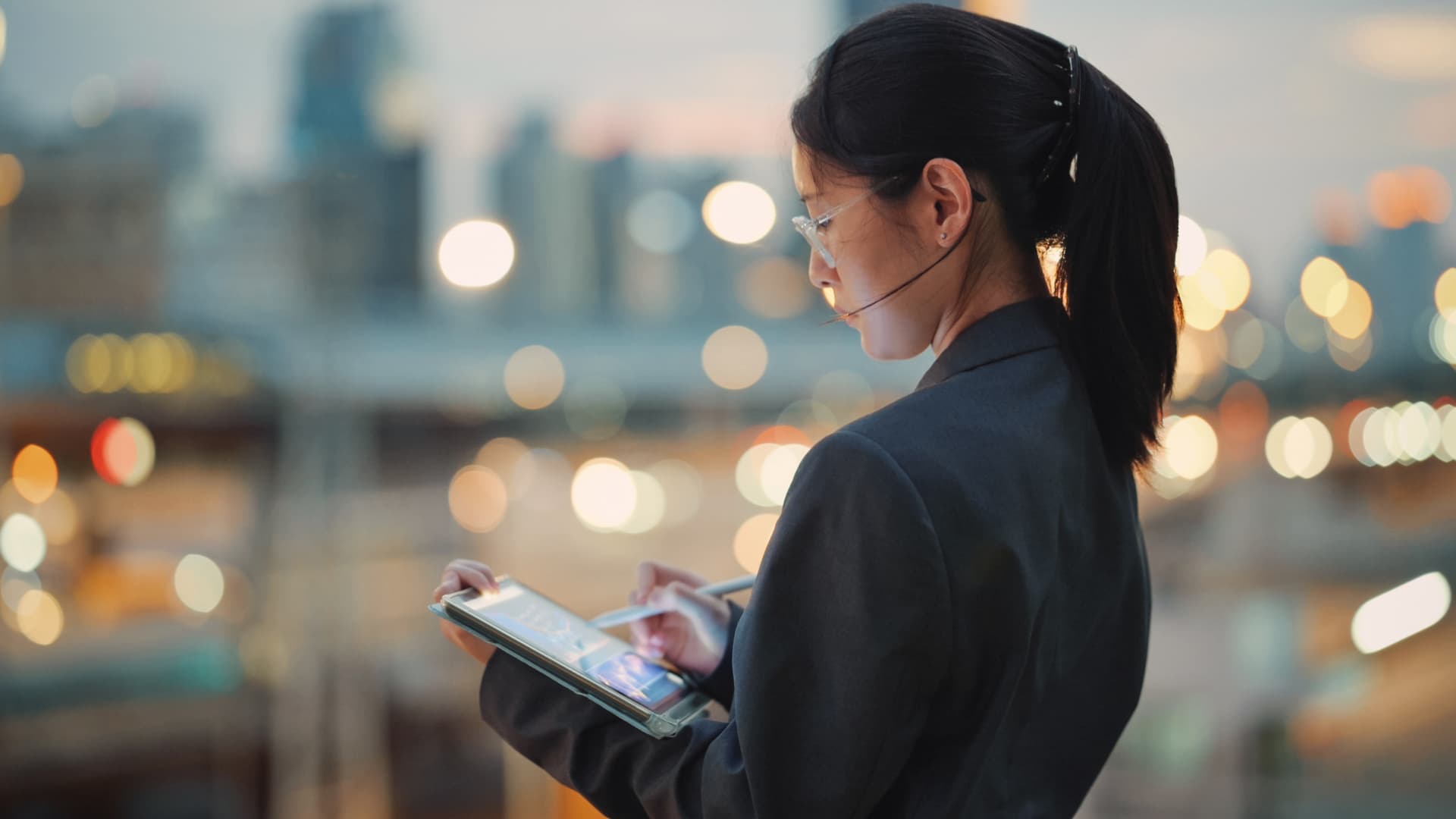 Mujer de negocios utilizando su tablet con la ciudad de fondo atardeciendo, luces de edificios y rascacielos.
