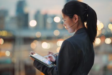 Mujer de negocios utilizando su tablet con la ciudad de fondo atardeciendo, luces de edificios y rascacielos.