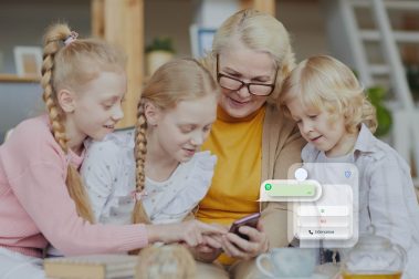 3 hermanos niños de pelo rubio junto a su abuela mirando el smartphone utilizando WhatsApp en casa.